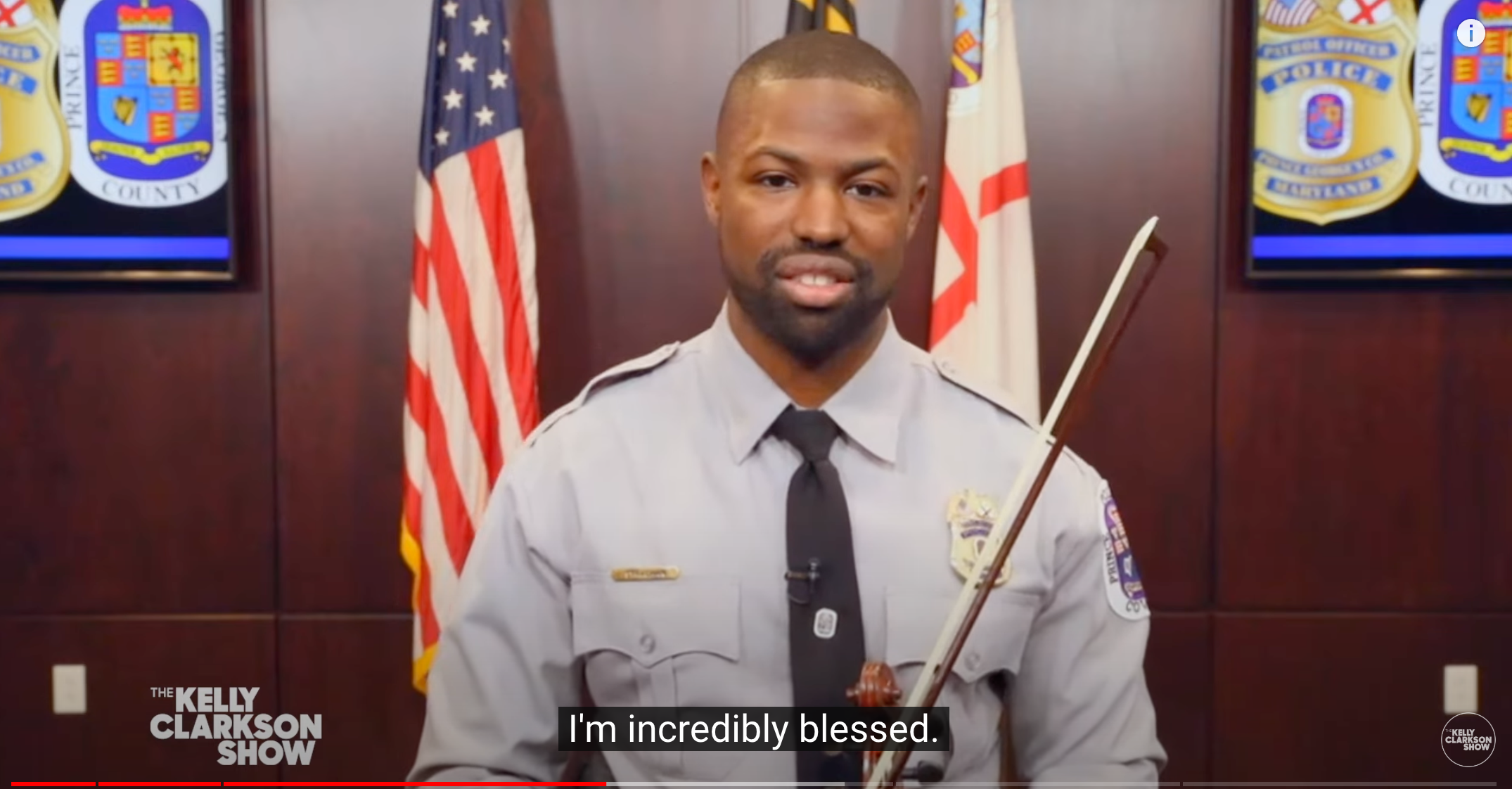 Alex Strachen appears on Kelly Clarkson show in May 2023, wearing police uniform and holding his violin with US flag behind his shoulder.